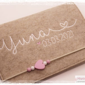 May include: A beige felt pouch with a white embroidered name "Yuna" and the date "03.03.2021". A pink heart and a string of pink and white beads are attached to the pouch.