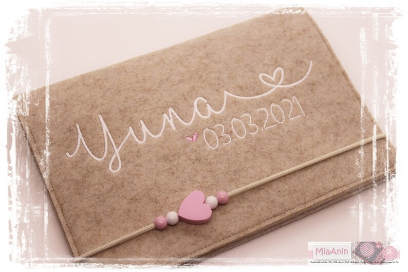 May include: A beige felt pouch with a white embroidered name "Yuna" and the date "03.03.2021". A pink heart and a string of pink and white beads are attached to the pouch.