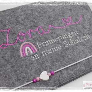 May include: A grey felt storage box with a pink rainbow and the text "Zora" and "Erinnerungen an meine Schulzeit" embroidered on it. A white string with pink beads and a white heart charm is attached to the box.
