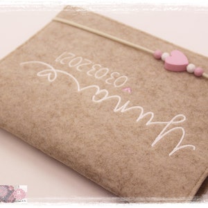 May include: A brown felt notebook with a white embroidered design that says "Mum" and "03.03.2021". A pink heart and a string of beads with pink hearts are attached to the notebook.