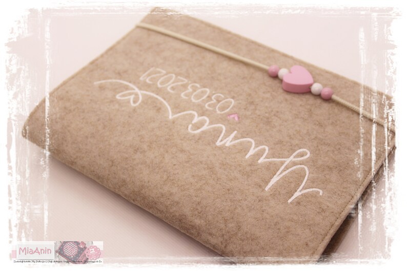 May include: A brown felt notebook with a white embroidered design that says "Mum" and "03.03.2021". A pink heart and a string of beads with pink hearts are attached to the notebook.