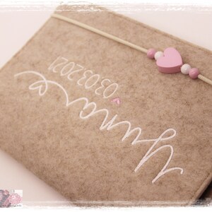 May include: A beige felt book with white embroidery that reads "Love" and "12.02.2020". A pink heart and white beads are attached to a white string that wraps around the book.