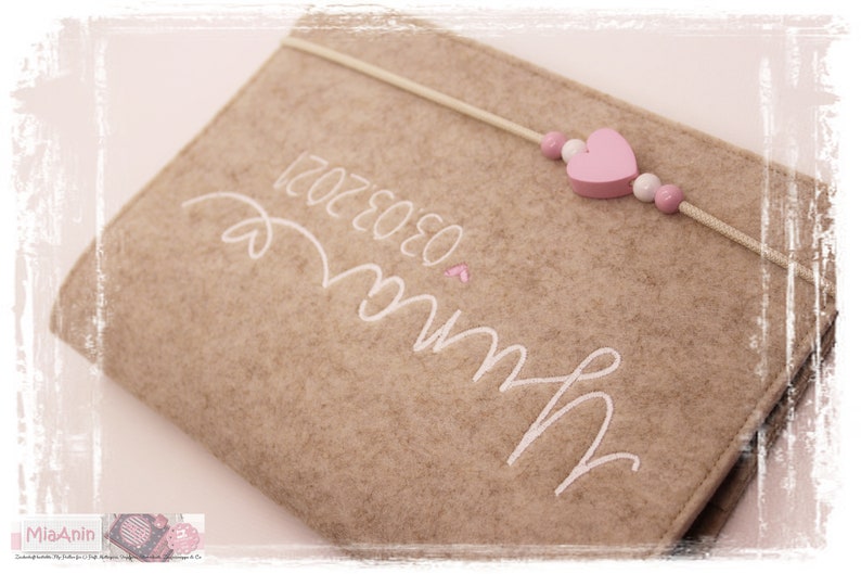 May include: A beige felt book with white embroidery that reads "Love" and "12.02.2020". A pink heart and white beads are attached to a white string that wraps around the book.