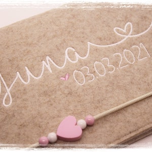 May include: A beige felt keepsake box with a white embroidered name "Yuna" and the date "03.03.2021". A pink wooden heart charm is attached to a white cord.