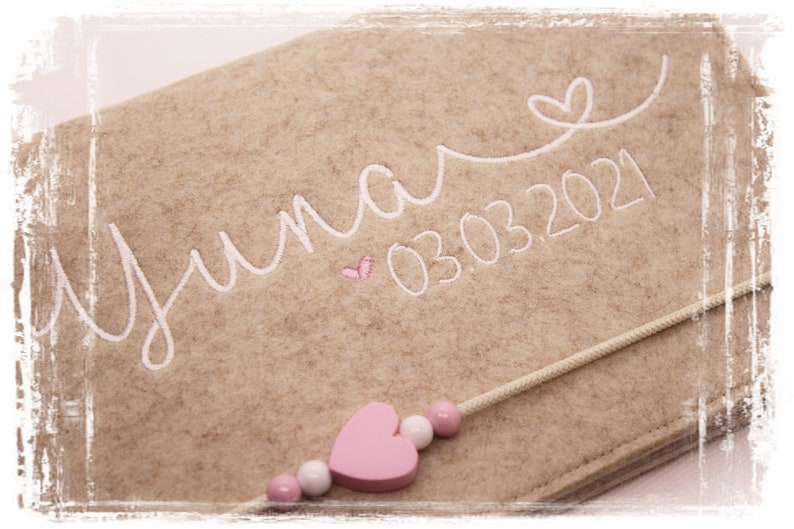 May include: A beige felt keepsake box with a white embroidered name "Yuna" and the date "03.03.2021". A pink wooden heart charm is attached to a white cord.