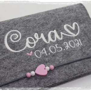 Pu&ograve; includere: Borsa in feltro grigio con un nome ricamato in bianco "Cora" e la data "04.05.2021". La borsa ha una cerniera e un ciondolo a forma di cuore rosa con perline rosa e bianche.