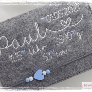 May include: Grey felt baby keepsake with white embroidery that reads "Paul" 01.05.2021 11:15 Uhr 3890 g 53 cm. The keepsake has a zipper closure and is decorated with blue and white heart-shaped embellishments.