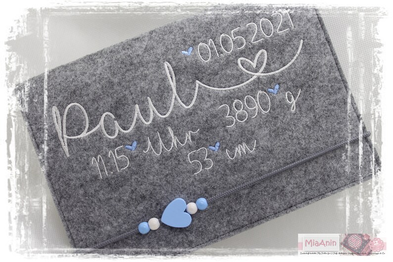 May include: Grey felt baby keepsake with white embroidery that reads "Paul" 01.05.2021 11:15 Uhr 3890 g 53 cm. The keepsake has a zipper closure and is decorated with blue and white heart-shaped embellishments.