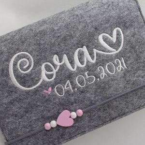 Pu&ograve; includere: Libro per neonati in feltro grigio con ricamo bianco che dice "Cora" con un cuore e la data "04.05.2021". Il libro ha un cuore rosa e perline bianche sul fondo.