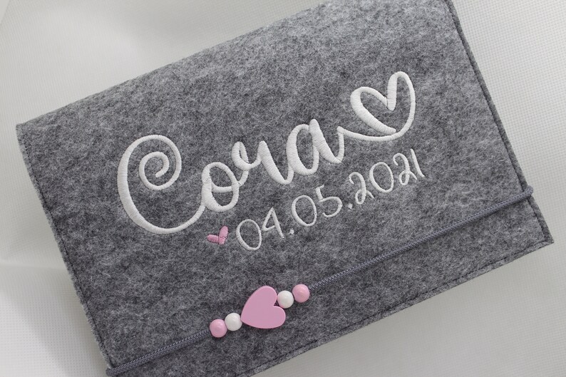 Pu&ograve; includere: Libro per neonati in feltro grigio con ricamo bianco che dice "Cora" con un cuore e la data "04.05.2021". Il libro ha un cuore rosa e perline bianche sul fondo.