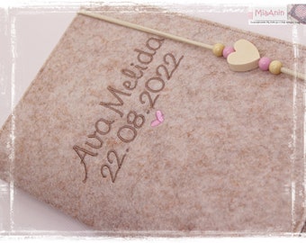 Può includere: Un libro di ricordi per neonati in feltro beige con il nome "Alva Melina" e la data "22.08.2022" ricamate sulla copertina. Un cuore rosa e un cuore bianco con una collana di perline sono attaccati alla copertina.