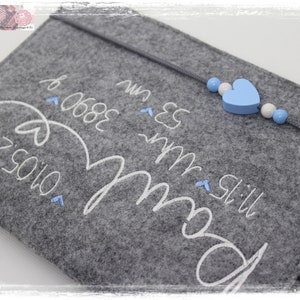 May include: Grey felt pouch with white embroidery that reads "Still my boy" and "06.08.2023" with a blue heart and white beads attached to a string.