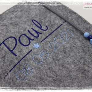 May include: Grey felt baby blanket with blue embroidery that reads "Paul 08.05.2021". A blue star and a blue wooden star with beads are attached to the blanket.