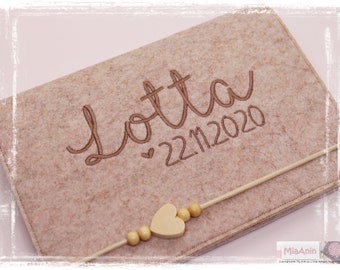 Può includere: Una scatola portaoggetti in feltro beige con il nome "Lotta" ricamato in filo marrone e la data "22.11.2020" ricamata sotto. Un ciondolo a forma di cuore in legno con una corda beige di perline di legno è attaccato alla scatola.