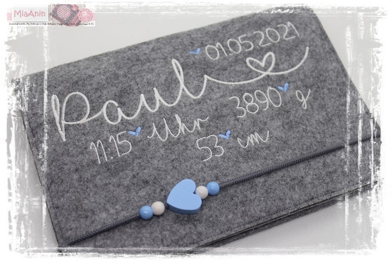May include: Grey felt baby keepsake with white embroidery that reads "Paul 01.05.2021 11:15 Uhr 3890 g 53 cm" with a blue heart and beads.