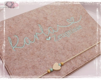 Può includere: Un portadocumenti in feltro marrone chiaro con il nome "Karla" e la parola "Zeugnisse" ricamati in azzurro. Presenta una corda decorativa con un cuore e perline di legno.