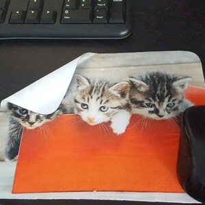 May include: A mouse pad with a picture of three kittens in a red box. The kittens are gray and white with blue eyes. The mouse pad is on a black surface.