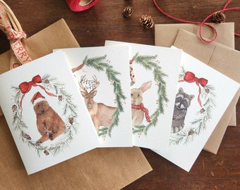 Pode incluir: Conjunto de quatro cartões de Natal em aquarela com animais da floresta em grinaldas. Cada cartão apresenta um animal diferente: um urso, um veado, um coelho e um guaxinim. Os cartões têm laços vermelhos e detalhes de pinhas, com envelopes de papel castanho.