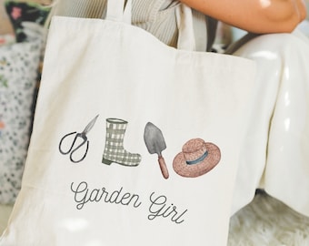 May include: Cream-colored tote bag featuring watercolor illustrations of gardening tools: scissors, a plaid rain boot, a trowel, and a straw hat. The words "Garden Girl" are written in cursive below the illustrations. The bag has two cream-colored handles.