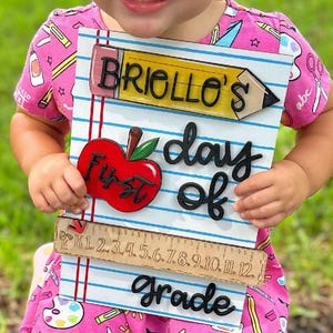 May include: A pink outfit with school-themed patterns, including pencils and scissors. The outfit is paired with pink sneakers. A wooden sign reading "Briollo's day of first grade" is held in front of the outfit.