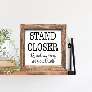 May include: A wooden sign with the text "STAND CLOSER it's not as long as you think" in black lettering. The sign is on a wooden shelf with a green plant hanging down.