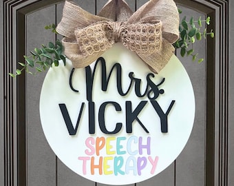 May include: A white round door hanger with a brown burlap bow and green leaves. The text "Mrs. Vicky Speech Therapy" is written in black and colorful letters.