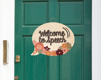 May include: A wooden speech bubble door hanger with the text "Welcome to Speech" in brown lettering. The speech bubble is decorated with a floral pattern in shades of orange, pink, and green.
