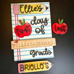 May include: A wooden school-themed sign with the words "Ellie's" and "Briollo's" on pencil-shaped cutouts. The sign also features a red apple with "First" and "Last" on them, a ruler, and the words "day of grade".