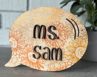 May include: Wooden speech bubble with a sunflower pattern and the text "ms sam" in brown letters.