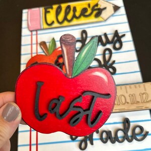May include: A wooden apple with the word "Last" in black script is held in front of a school-themed background. The background includes a pencil, the name "Ellie's", and the words "Last Day of Grade". A wooden ruler is also visible.