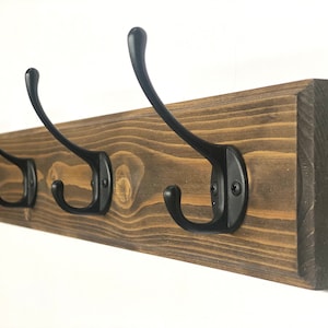 May include: A wooden coat rack with five black metal hooks. The rack is stained a dark brown color and has a rustic look.