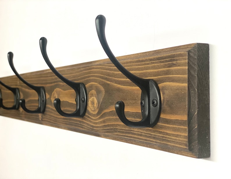 May include: A wooden coat rack with five black metal hooks. The rack is stained a dark brown color and has a rustic look.