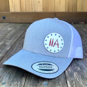 May include: A light blue snapback hat with a white mesh back. The hat features a circular patch with the letters "IIA" and a rifle graphic, surrounded by blue stars. The hat has a curved brim and the text "SNAPBACK".