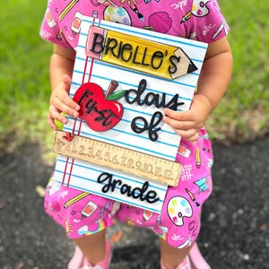May include: A wooden sign with the text "BRIELLE'S First days of Grade" and a red apple. The sign also features a pencil, ruler, and blue lined paper design. The sign is held by a person wearing a pink outfit.