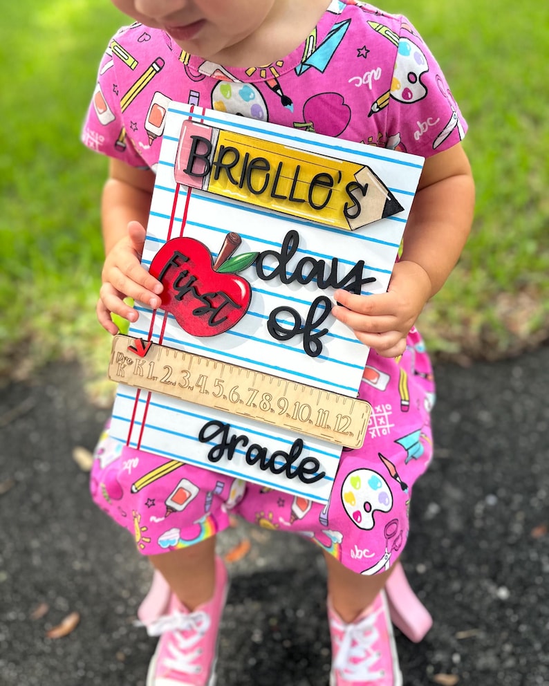 May include: A wooden sign with the text "BRIELLE'S First days of Grade" and a red apple. The sign also features a pencil, ruler, and blue lined paper design. The sign is held by a person wearing a pink outfit.