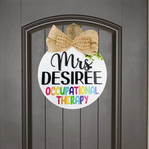 May include: A white round door hanger with the text "Mrs. DESIREE OCCUPATIONAL THERAPY" in black and rainbow letters. The door hanger is decorated with a brown burlap bow and green leaves.