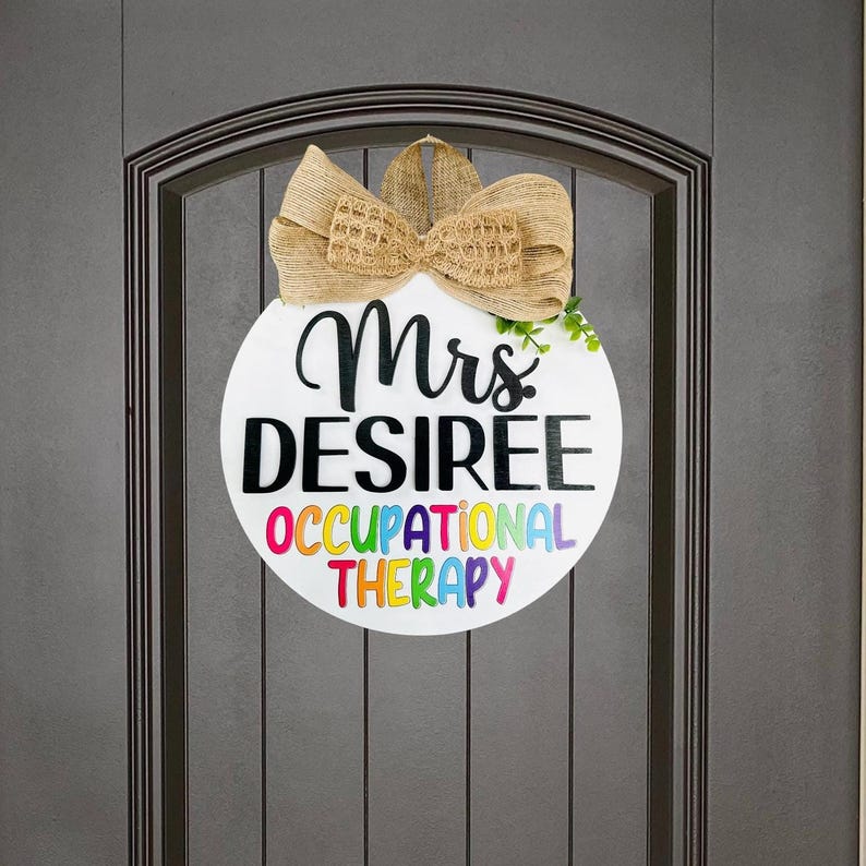May include: A white round door hanger with the text "Mrs. DESIREE OCCUPATIONAL THERAPY" in black and rainbow letters. The door hanger is decorated with a brown burlap bow and green leaves.