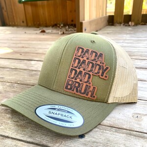 May include: A green and beige trucker hat with a brown leather patch that says "Dada, Daddy, Dad,  Bruh!" The hat has a mesh back and a snap closure. The hat is made by Yupoong and is an authentic snapback.