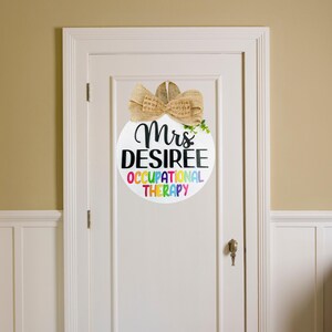 May include: A white round door hanger with a burlap bow. The text on the door hanger reads "Mrs. DESIREE OCCUPATIONAL THERAPY" in black text with rainbow colored letters for "OCCUPATIONAL THERAPY".