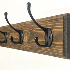 May include: A wooden coat rack with five black metal hooks. The rack is stained a dark brown color and has a rustic look.