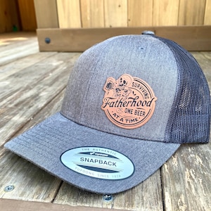 May include: A gray and black mesh baseball cap with a brown leather patch that says "Surviving Fatherhood & One Beer At A Time". The patch features a cartoon skeleton holding a beer mug. The cap has a white label that says "The Authentic YUPOONG Snapback YUPOONG Since 1974".