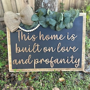 May include: A black wooden sign with a rustic wood frame. The sign has a white pearl garland and a burlap bow at the top. The sign says "This home is built on love and profanity" in gold lettering.