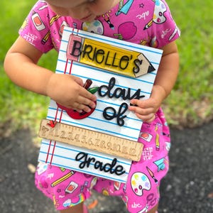May include: A wooden sign with the text "Briollo's days of grade" in black, with a pencil and apple design. The sign has a ruler at the bottom and is held by a person wearing a pink dress with school-themed patterns.