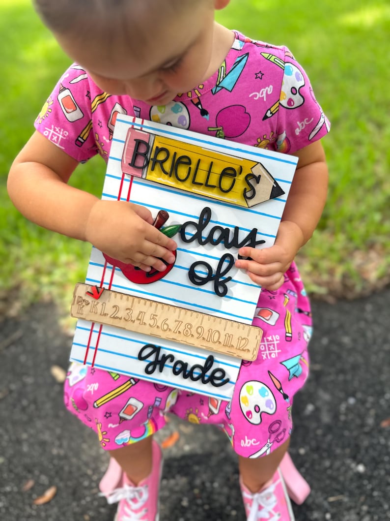 May include: A wooden sign with the text "Briollo's days of grade" in black, with a pencil and apple design. The sign has a ruler at the bottom and is held by a person wearing a pink dress with school-themed patterns.