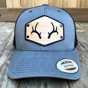 May include: A gray and black trucker hat with a wooden patch featuring a pair of antlers. The patch has black rivets around the edge. The hat has a black mesh back and a black snap closure. The hat has a white label on the back that says "The Authentic Yupoong Snapback Yupoong Since 1974."