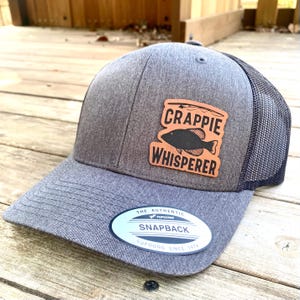 May include: A gray and black mesh trucker hat with a brown leather patch that says "Crappie Whisperer" and a black silhouette of a fish. The hat also has a white oval sticker that says "The Authentic Yupoong Snapback Yupoong Since 1974".