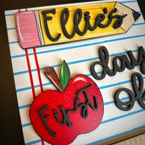 May include: A wooden sign with a school theme. The sign features a yellow pencil with the name "Ellie's" in black script. A red apple with the words "First day of" in black script is also present. The background is white with blue and red lines.
