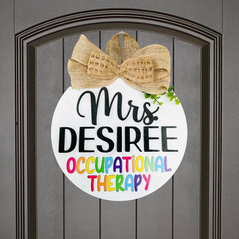 May include: A white round door hanger with a brown burlap bow. The text on the door hanger reads "Mrs. DESIREE OCCUPATIONAL THERAPY" with the words "OCCUPATIONAL THERAPY" in rainbow colors.