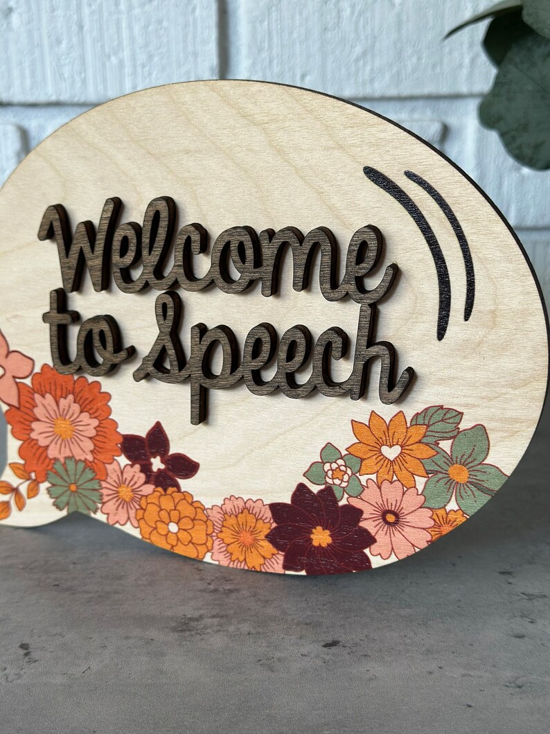 May include: A wooden sign in the shape of a speech bubble with the text "Welcome to Speech" in brown lettering. The sign is decorated with a border of colorful flowers.
