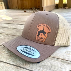 May include: Brown and tan baseball cap with a brown leather patch that says "Mind Your Business My Wife Is Expensive". The cap has a mesh back and a logo that says "The Authentic Yupoong Snapback Since 1974".